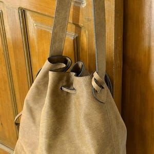 May include: A tan bucket bag with a soft, matte texture and a drawstring closure. The bag features a single long shoulder strap that attaches to the sides, creating a gathered opening at the top. The material has a consistent, muted brown tone.
