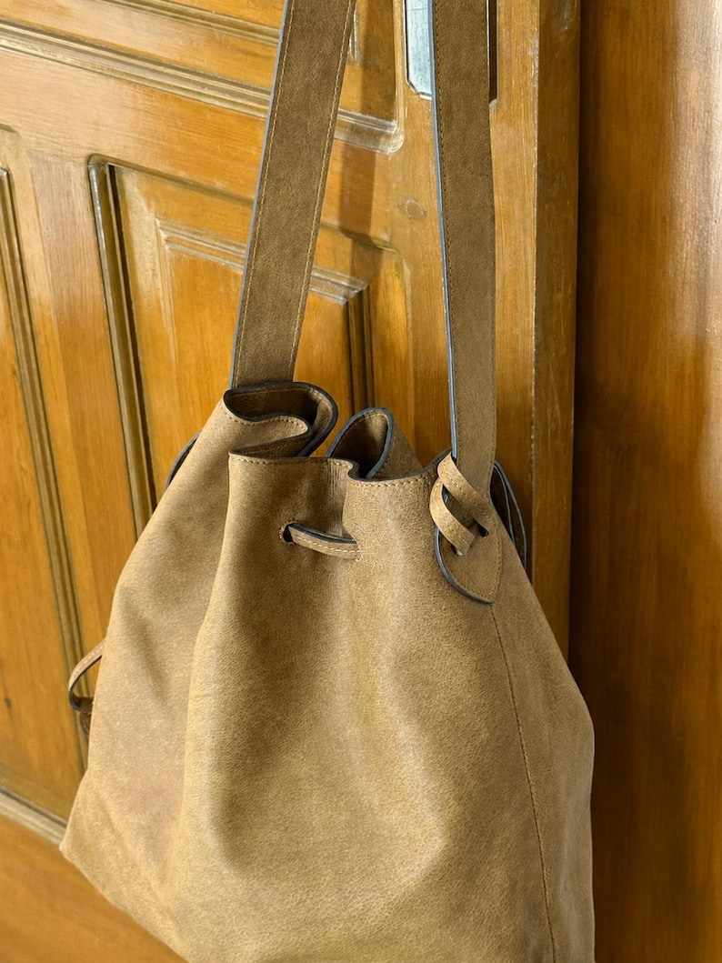 May include: A tan bucket bag with a soft, matte texture and a drawstring closure. The bag features a single long shoulder strap that attaches to the sides, creating a gathered opening at the top. The material has a consistent, muted brown tone.