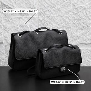 Black top grain leather shoulder bags in two sizes displayed on a dark tabletop against a textured stone wall. Size dimensions are highlighted: Large bag measures 15.4" W x 9.8" H x 4.7" D, and small bag measures 12.6" W x 7.5" H x 4.3" D. Features a minimalist silhouette with silver eyelets and structured flap design.