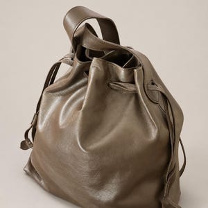 May include: A taupe-colored leather bucket bag featuring a drawstring closure with matching leather cords. The bag has a single top handle and a soft, slouchy structure with visible folds in the material, creating a rounded, voluminous silhouette.