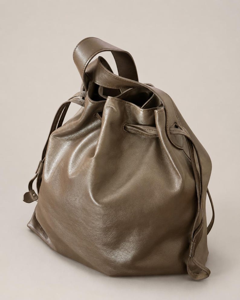 May include: A taupe-colored leather bucket bag featuring a drawstring closure with matching leather cords. The bag has a single top handle and a soft, slouchy structure with visible folds in the material, creating a rounded, voluminous silhouette.