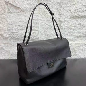 Black leather handbag shown from an angled view with a structured rectangular shape, long shoulder strap, flap closure, and silver hardware details. This elegant purse offers a sophisticated look with durable construction and a classic minimalist style.
