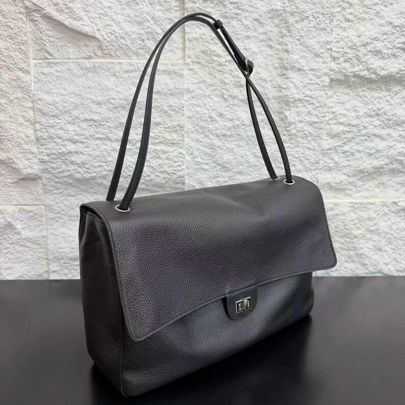 Black leather handbag shown from an angled view with a structured rectangular shape, long shoulder strap, flap closure, and silver hardware details. This elegant purse offers a sophisticated look with durable construction and a classic minimalist style.