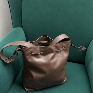 May include: A taupe-colored leather bucket bag featuring a drawstring closure, a single shoulder strap, and a soft, slouchy silhouette. The bag has a smooth, matte finish and includes metal eyelets for the drawstring cord to pass through.