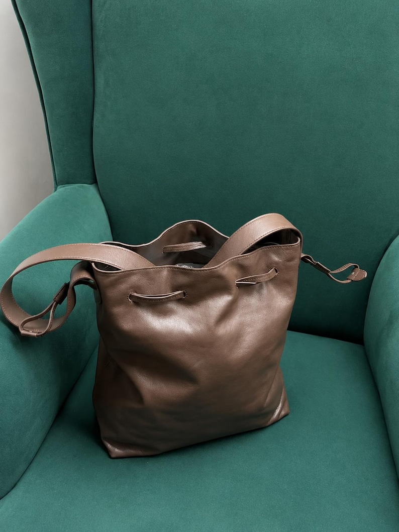 May include: A taupe-colored leather bucket bag featuring a drawstring closure, a single shoulder strap, and a soft, slouchy silhouette. The bag has a smooth, matte finish and includes metal eyelets for the drawstring cord to pass through.