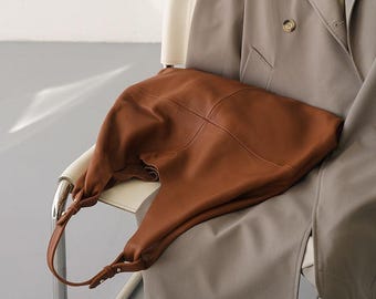 May include: A brown leather shoulder bag with a soft, slouchy design. The bag has a single strap and is resting on a white chair. The bag is made of a smooth leather material and has a simple, elegant style.