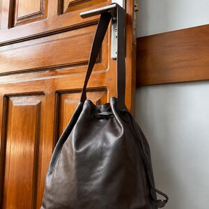 May include: A dark brown leather drawstring bucket bag with a single long shoulder strap. The bag features a smooth, matte finish and a cinched top closure secured by a matching leather drawstring cord that hangs down the side of the bag.