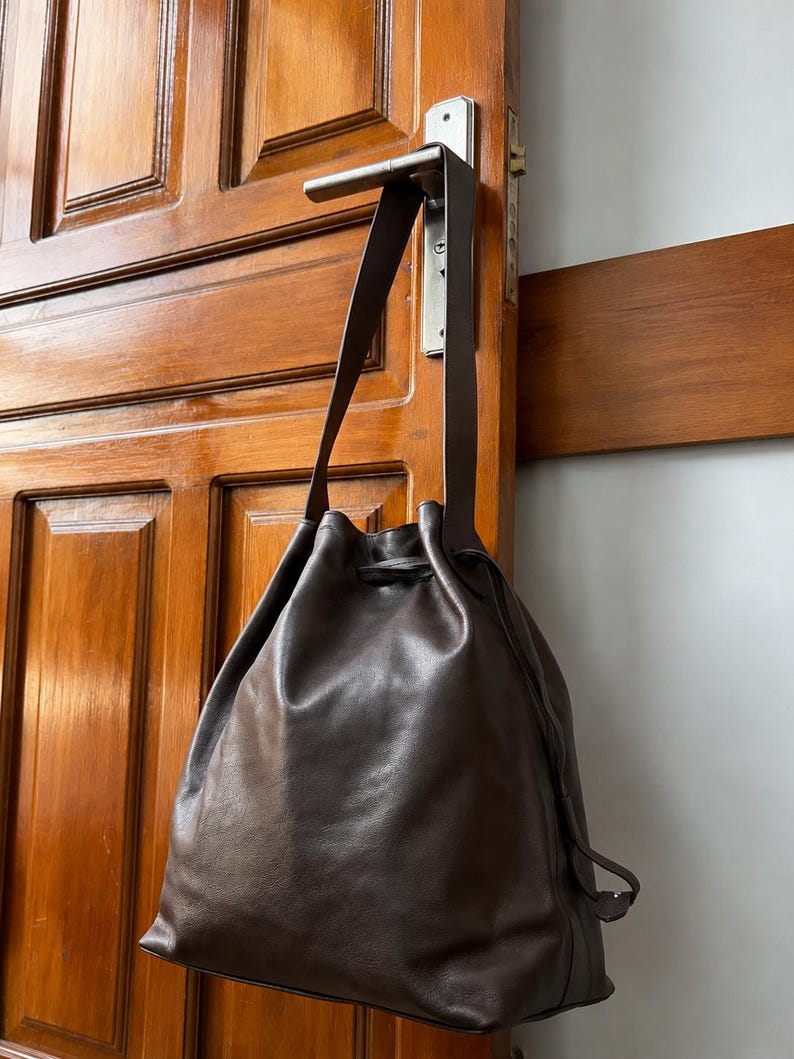 May include: A dark brown leather drawstring bucket bag with a single long shoulder strap. The bag features a smooth, matte finish and a cinched top closure secured by a matching leather drawstring cord that hangs down the side of the bag.
