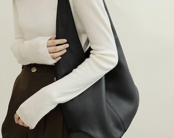 May include: A black leather shoulder bag with a rounded bottom and a single strap. The bag is held against a person wearing a white turtleneck sweater and brown pants.