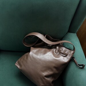May include: A brown leather bucket bag featuring a smooth, soft texture, a single shoulder strap, and a drawstring closure mechanism with thin leather ties that cinch the top opening of the bag to secure its contents.