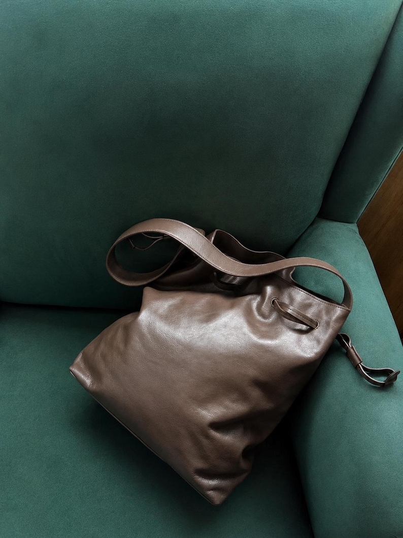 May include: A brown leather bucket bag featuring a smooth, soft texture, a single shoulder strap, and a drawstring closure mechanism with thin leather ties that cinch the top opening of the bag to secure its contents.