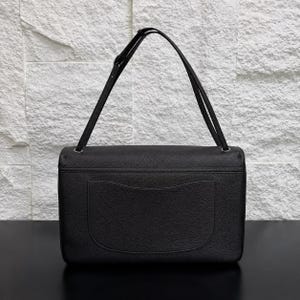 Back view of a black top grain leather shoulder bag featuring a clean, minimalist design with a spacious exterior slip pocket and adjustable shoulder strap. The structured silhouette and textured leather finish are showcased against a textured stone backdrop, highlighting its practical storage and timeless everyday style.