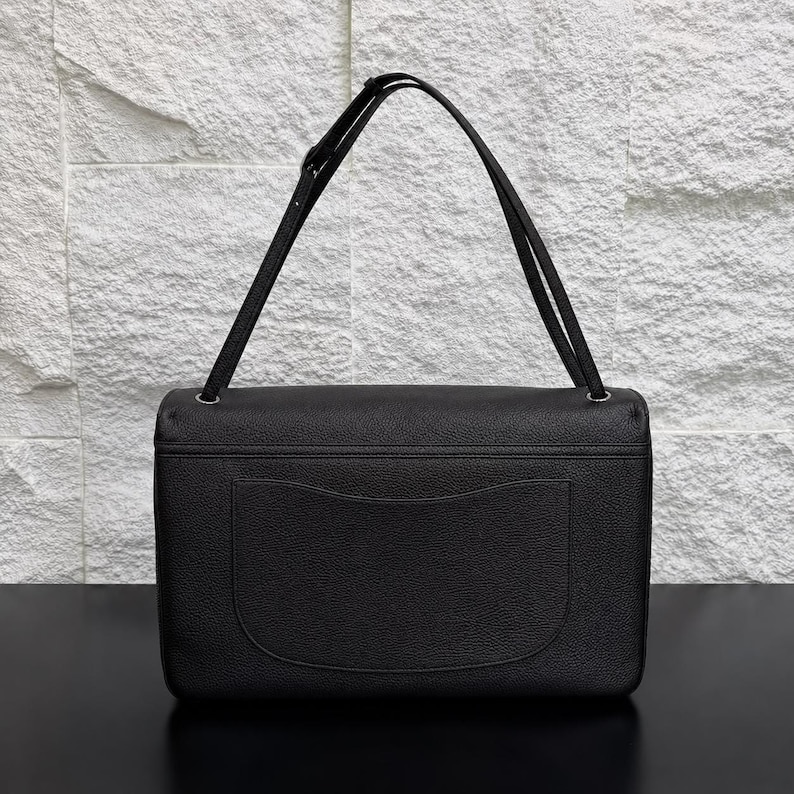 Back view of a black top grain leather shoulder bag featuring a clean, minimalist design with a spacious exterior slip pocket and adjustable shoulder strap. The structured silhouette and textured leather finish are showcased against a textured stone backdrop, highlighting its practical storage and timeless everyday style.