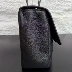 Side profile of a black leather handbag displaying its structured shape, sturdy construction, and textured leather surface. The elegant design includes a spacious body, refined stitching, and a comfortable shoulder strap, making it a versatile accessory for daily use.