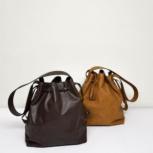 May include: Two bucket bags with drawstring closures and shoulder straps. One bag is dark brown with a smooth finish, and the other is a lighter tan color with a textured, matte finish. Both bags feature a cinched top design and long straps.
