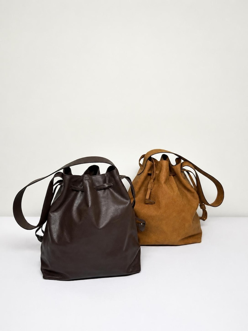 May include: Two bucket bags with drawstring closures and shoulder straps. One bag is dark brown with a smooth finish, and the other is a lighter tan color with a textured, matte finish. Both bags feature a cinched top design and long straps.