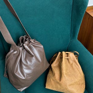 May include: Two drawstring bucket bags. One bag is a smooth, glossy taupe leather with a long shoulder strap. The second bag is a textured, matte tan suede. Both bags feature a cinched top opening with matching leather drawstring cords.