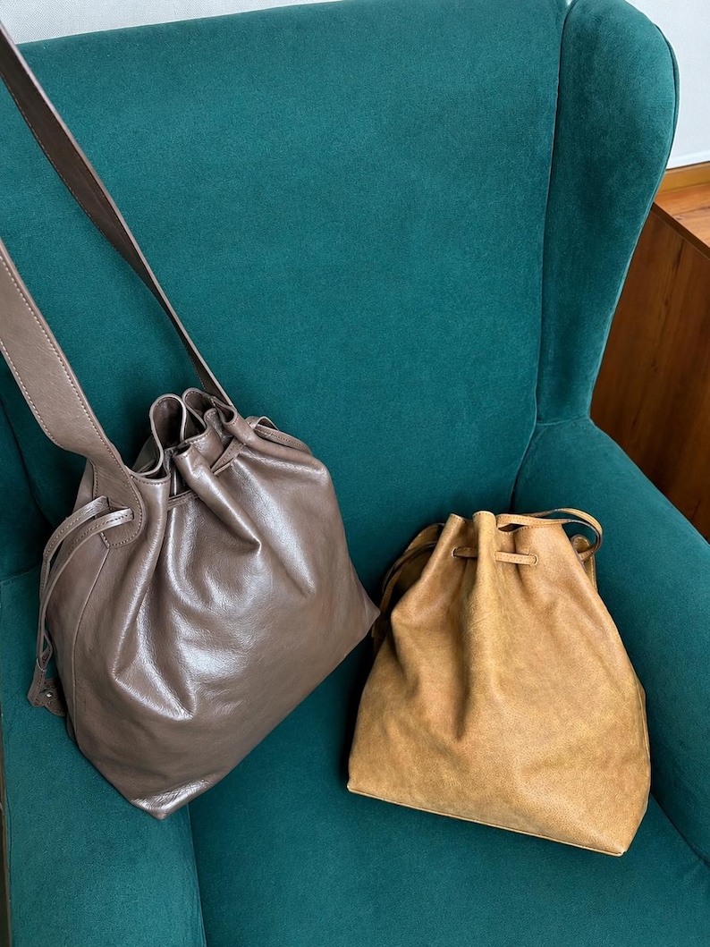 May include: Two drawstring bucket bags. One bag is a smooth, glossy taupe leather with a long shoulder strap. The second bag is a textured, matte tan suede. Both bags feature a cinched top opening with matching leather drawstring cords.