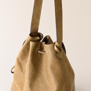 May include: A tan bucket bag with a textured finish, featuring a drawstring closure threaded through metal eyelets and a single long shoulder strap attached to the sides. The bag has a soft, slouchy structure and a consistent earthy color tone.