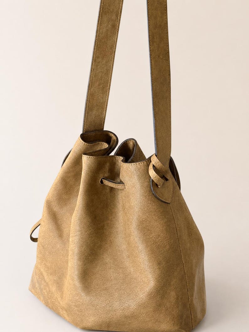 May include: A tan bucket bag with a textured finish, featuring a drawstring closure threaded through metal eyelets and a single long shoulder strap attached to the sides. The bag has a soft, slouchy structure and a consistent earthy color tone.