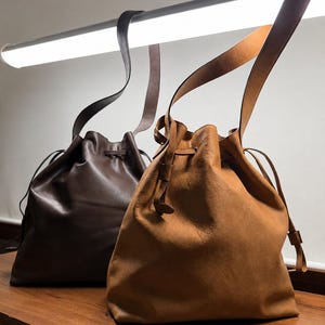May include: Two leather bucket bags with drawstring closures and long shoulder straps. One bag is a deep dark brown with a smooth finish, while the other is a lighter tan color with a slightly textured, matte appearance. Both bags feature cinched tops.