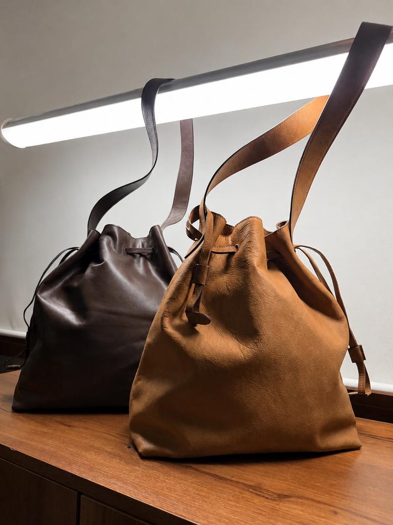 May include: Two leather bucket bags with drawstring closures and long shoulder straps. One bag is a deep dark brown with a smooth finish, while the other is a lighter tan color with a slightly textured, matte appearance. Both bags feature cinched tops.