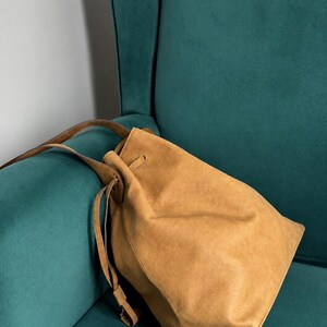 May include: A tan suede bucket bag featuring a drawstring closure with matching thin leather straps. The bag has a soft, unstructured silhouette and a smooth, matte finish, showcasing a simple design with a single main compartment and long handles.