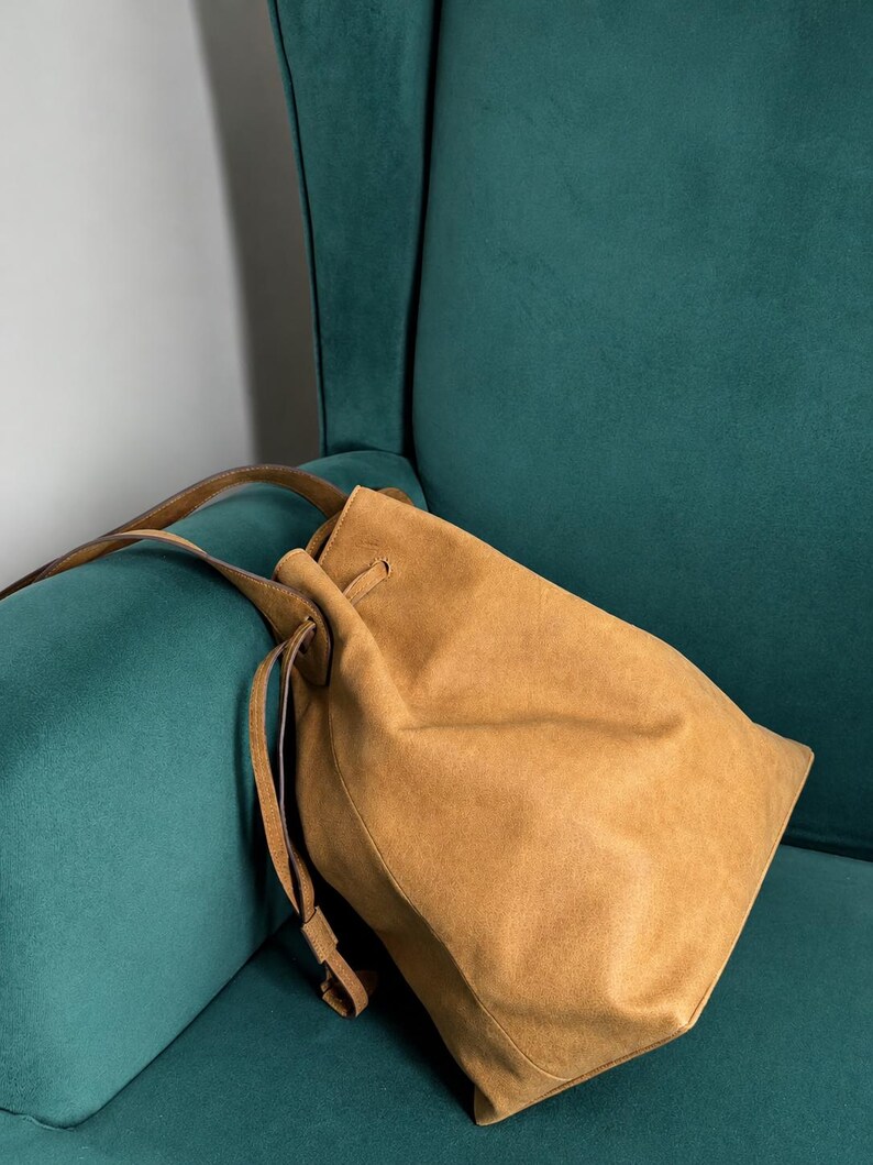 May include: A tan suede bucket bag featuring a drawstring closure with matching thin leather straps. The bag has a soft, unstructured silhouette and a smooth, matte finish, showcasing a simple design with a single main compartment and long handles.