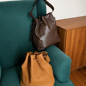 May include: Two drawstring bucket bags, one in a dark brown smooth finish and one in a tan suede-like texture. Both bags feature a single shoulder strap and a cinched top closure with matching drawstring cords, presented as a pair of accessories.