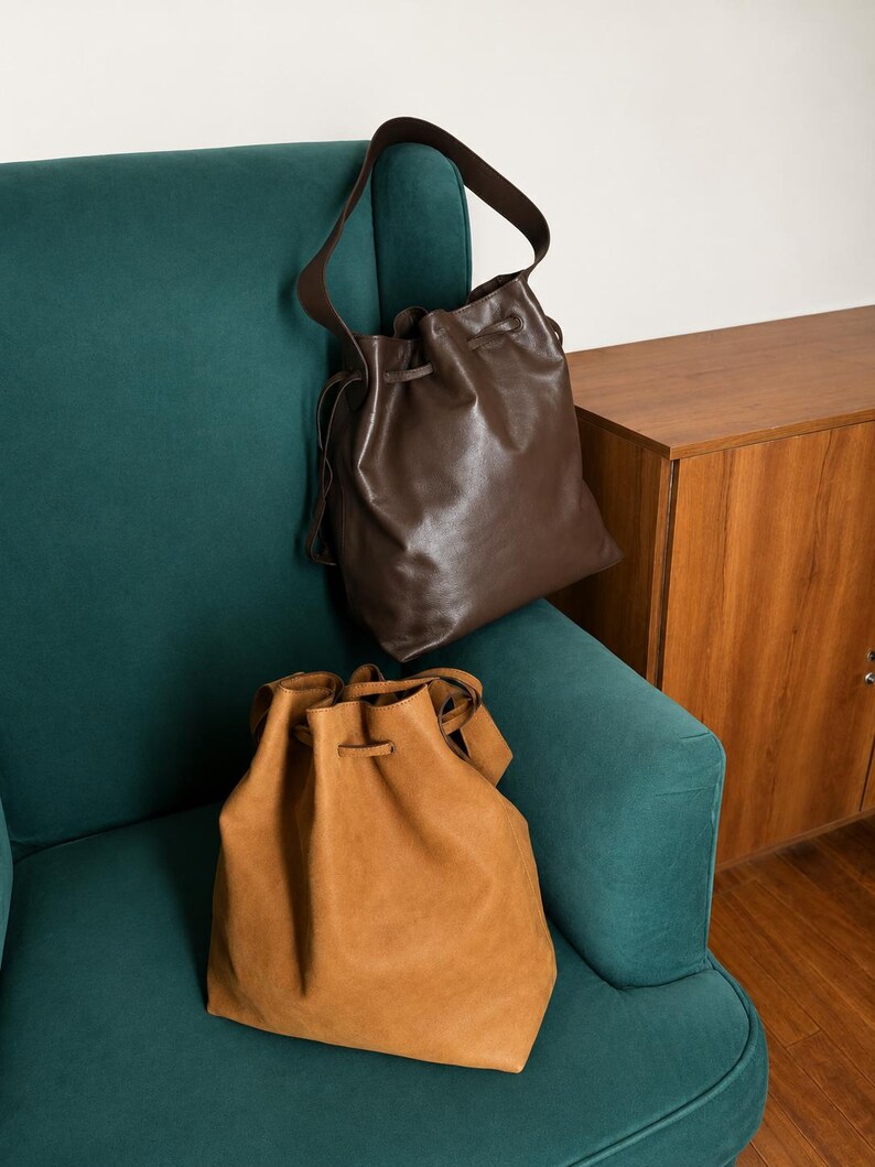 May include: Two drawstring bucket bags, one in a dark brown smooth finish and one in a tan suede-like texture. Both bags feature a single shoulder strap and a cinched top closure with matching drawstring cords, presented as a pair of accessories.