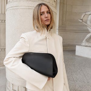 May include: A smooth black leather clutch bag with a rounded top edge and a structured silhouette, held under the arm. The bag features a minimalist design with a solid matte black finish and a clean, curved shape that contrasts with the cream jacket.