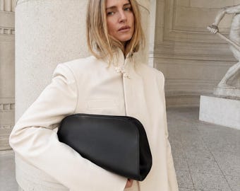May include: A smooth black leather clutch bag with a rounded top edge and a structured silhouette, held under the arm. The bag features a minimalist design with a solid matte black finish and a clean, curved shape that contrasts with the cream jacket.