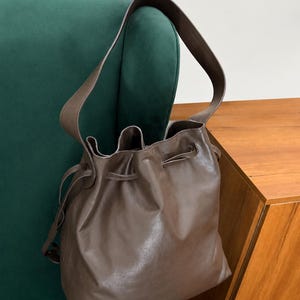 May include: A brown leather bucket bag featuring a smooth finish, a single shoulder strap, and a drawstring closure at the top. The bag has a soft, slouchy structure with visible folds around the opening and thin leather cords for cinching.