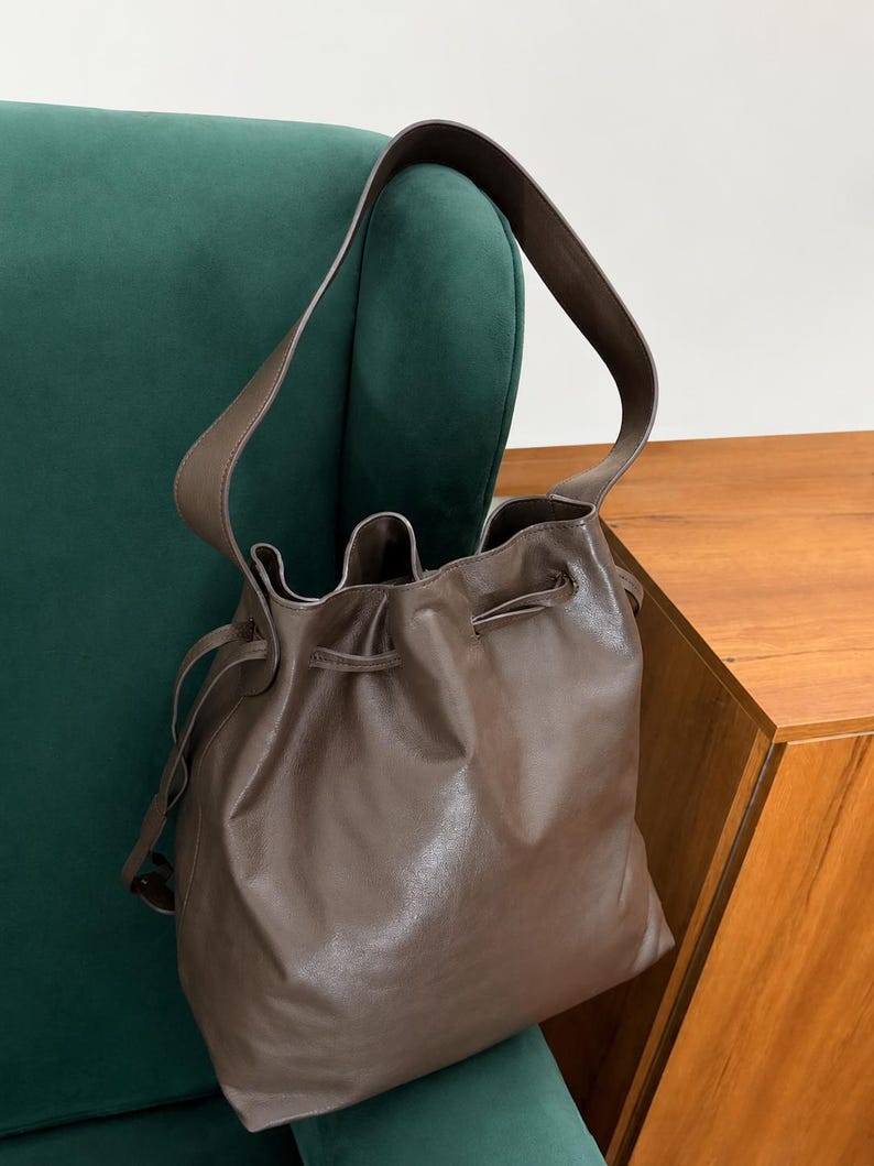 May include: A brown leather bucket bag featuring a smooth finish, a single shoulder strap, and a drawstring closure at the top. The bag has a soft, slouchy structure with visible folds around the opening and thin leather cords for cinching.