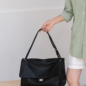 May include: A black leather shoulder bag with a textured surface, a large fold-over flap, and a silver-toned metal turn-lock closure. The bag features a long, adjustable shoulder strap attached with silver-toned hardware rings on both sides.