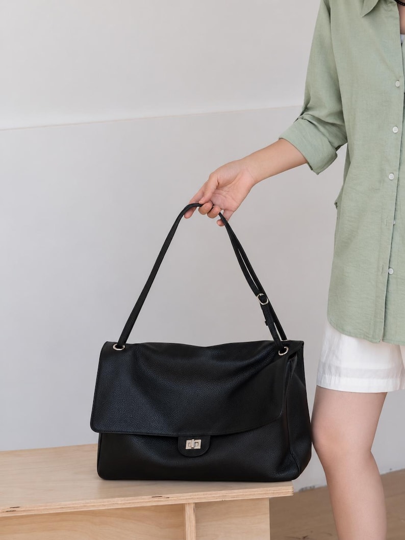 May include: A black leather shoulder bag with a textured surface, a large fold-over flap, and a silver-toned metal turn-lock closure. The bag features a long, adjustable shoulder strap attached with silver-toned hardware rings on both sides.