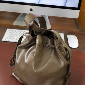 May include: A taupe-colored leather bucket bag featuring a single top handle and a drawstring closure mechanism. The bag has a smooth, slightly reflective surface with visible folds and a structured base, designed for carrying personal items.