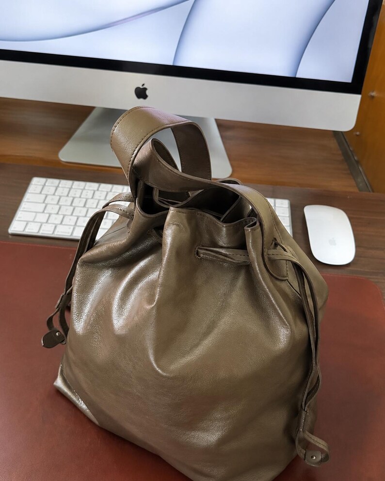 May include: A taupe-colored leather bucket bag featuring a single top handle and a drawstring closure mechanism. The bag has a smooth, slightly reflective surface with visible folds and a structured base, designed for carrying personal items.