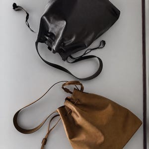 May include: Two drawstring bags, one in black and one in tan, featuring a smooth finish, a single shoulder strap, and a cinched top closure. The bags are positioned diagonally, showcasing the simple design and the long, thin leather-like straps.
