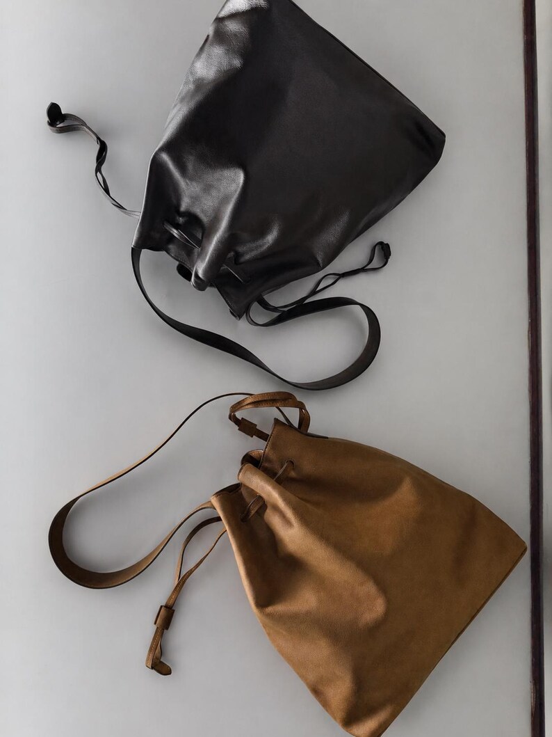 May include: Two drawstring bags, one in black and one in tan, featuring a smooth finish, a single shoulder strap, and a cinched top closure. The bags are positioned diagonally, showcasing the simple design and the long, thin leather-like straps.