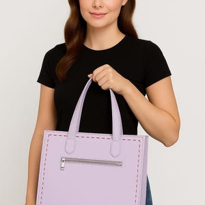 May include: A light purple tote bag with a rectangular shape and a zipper closure. The bag has two handles and red stitching details. The woman in the image is wearing a black t-shirt and blue jeans.