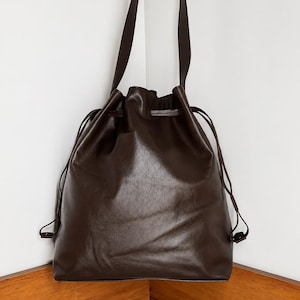 May include: A dark brown leather drawstring bag featuring a smooth, matte finish. The bag has a long shoulder strap and thin matching leather cords that cinch the top opening closed. The design is simple with a soft, flexible structure.