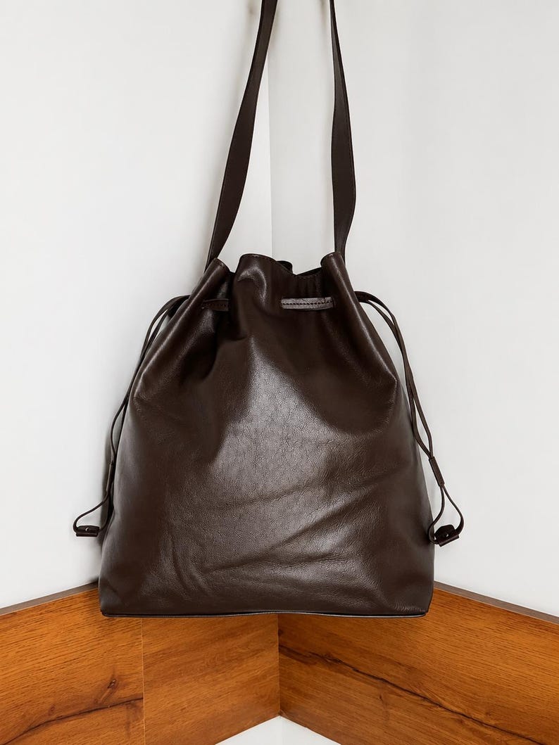 May include: A dark brown leather drawstring bag featuring a smooth, matte finish. The bag has a long shoulder strap and thin matching leather cords that cinch the top opening closed. The design is simple with a soft, flexible structure.