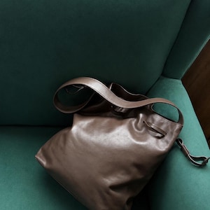 May include: A brown leather hobo bag featuring a slouchy, unstructured silhouette, a wide shoulder strap, and a drawstring closure mechanism with a thin leather cord detail. The bag has a smooth, matte finish and a soft, flexible construction.