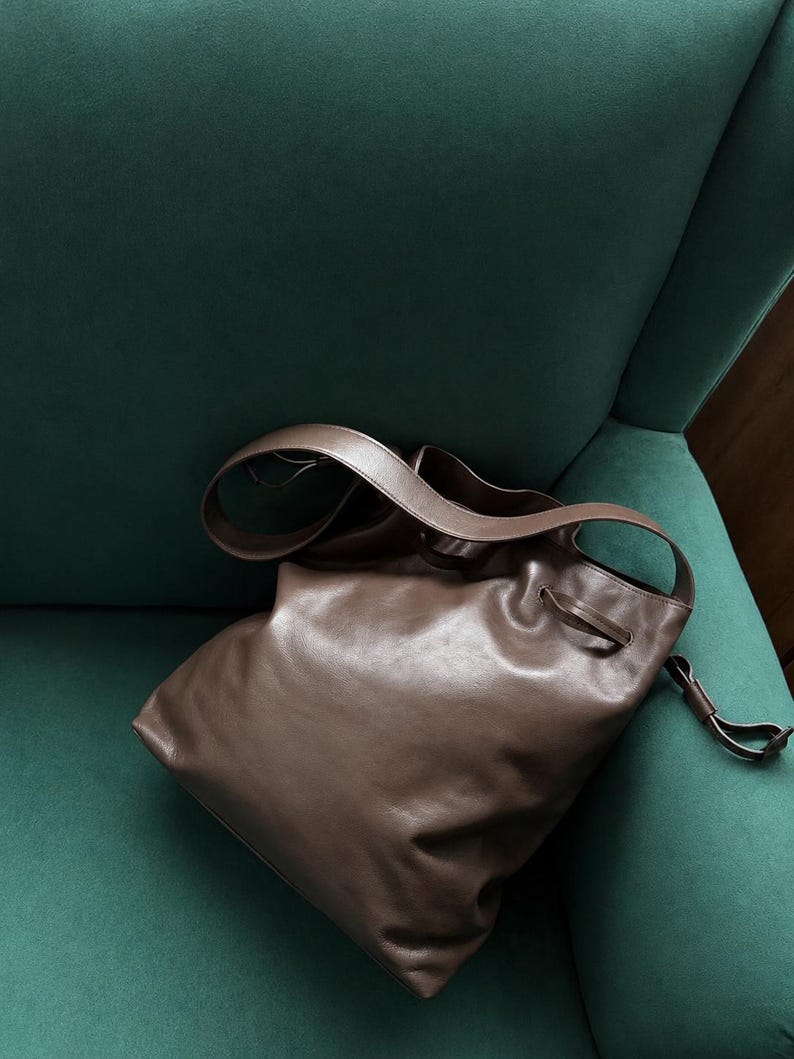 May include: A brown leather hobo bag featuring a slouchy, unstructured silhouette, a wide shoulder strap, and a drawstring closure mechanism with a thin leather cord detail. The bag has a smooth, matte finish and a soft, flexible construction.