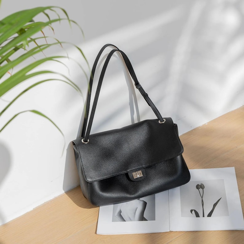 Black leather shoulder bag displayed on a wooden surface with a minimalist design. The handbag features a smooth flap closure, silver turn-lock buckle, adjustable strap, and spacious structured shape. A stylish everyday purse perfect for modern women, combining elegance, functionality, and timeless fashion.