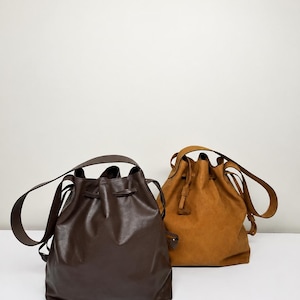 May include: Two bucket bags with drawstring closures and shoulder straps. One bag is dark brown with a smooth finish, and the other is a lighter tan color with a matte, textured finish. Both bags feature a rounded base and a cinched top design.