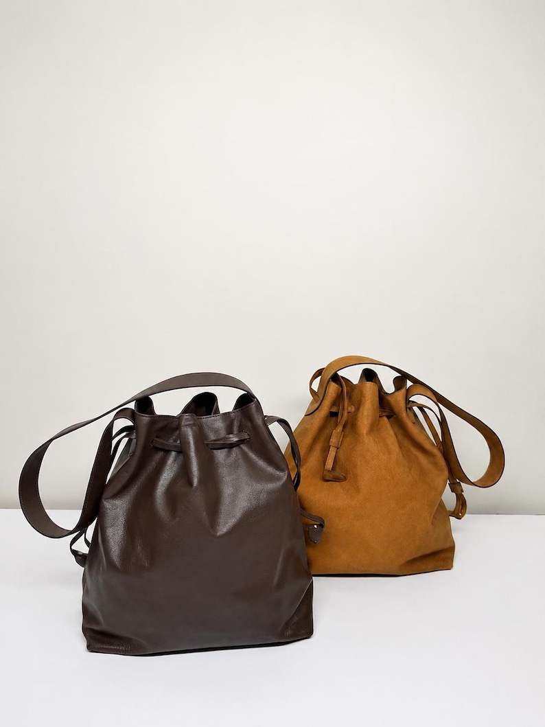 May include: Two bucket bags with drawstring closures and shoulder straps. One bag is dark brown with a smooth finish, and the other is a lighter tan color with a matte, textured finish. Both bags feature a rounded base and a cinched top design.