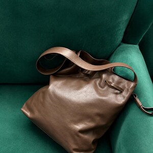May include: A brown leather bucket bag featuring a smooth, soft texture, a wide shoulder strap, and a drawstring closure mechanism with thin leather cords threaded through metal eyelets at the top opening of the bag.