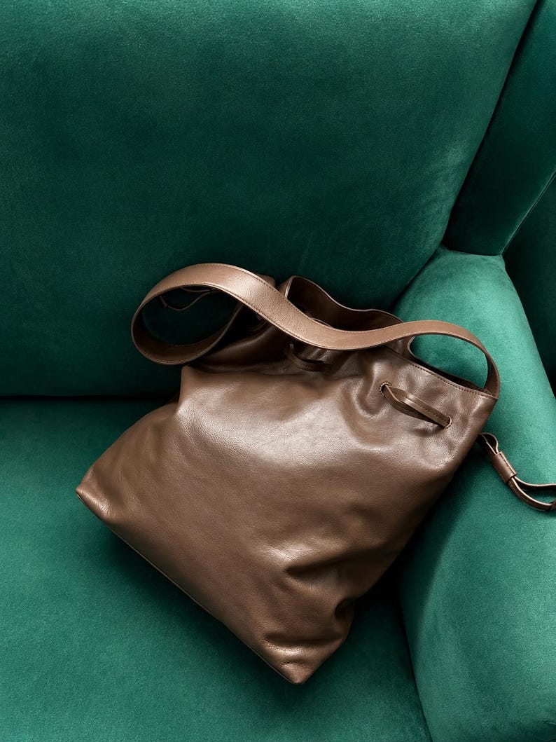 May include: A brown leather bucket bag featuring a smooth, soft texture, a wide shoulder strap, and a drawstring closure mechanism with thin leather cords threaded through metal eyelets at the top opening of the bag.